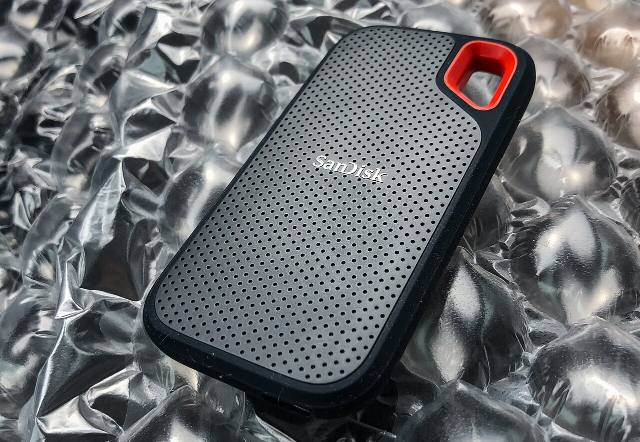 은색 완충재 위에 놓인 검은색 SanDisk 외장 SSD로, 모서리에 빨간 고리가 있어요.
